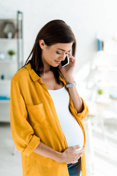 hermosa freelancer embarazada en camisa amarilla hablando en el teléfono inteligente en la oficina en casa
 - Foto, imagen