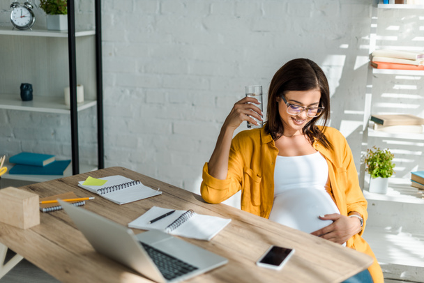 beautiful pregnant freelancer holding glass of water in office with home office - Photo, Image