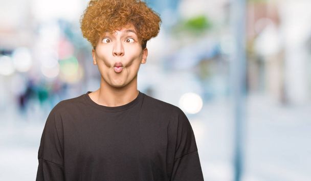 Young handsome man with afro hair wearing black t-shirt making fish face with lips, crazy and comical gesture. Funny expression. - Foto, imagen