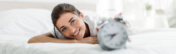 selective focus of happy girl lying with alarm clock on bed in the morning - 写真・画像