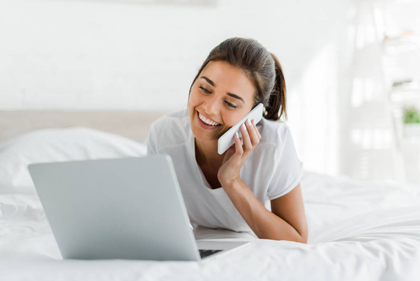 attractive smiling girl talking on smartphone and using laptop in bed in the morning  - Photo, Image