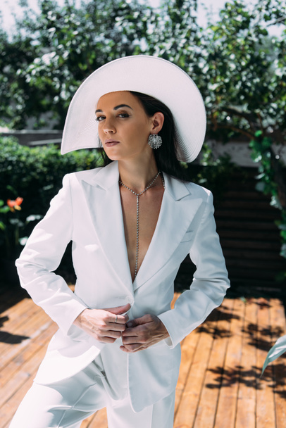 attractive woman in white suit and hat posing and looking at camera outside  - Foto, Imagem