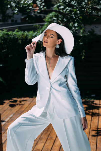 attractive woman in white suit and hat posing with closed eyes outside  - Fotó, kép