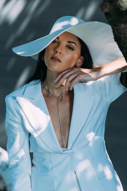 attractive woman in white suit and hat posing and looking at camera outside  - Foto, Imagen