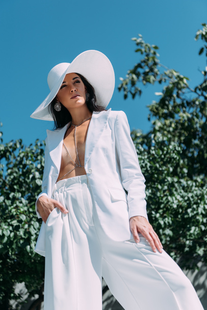 low angle view of attractive woman in white suit and hat posing outside  - Φωτογραφία, εικόνα