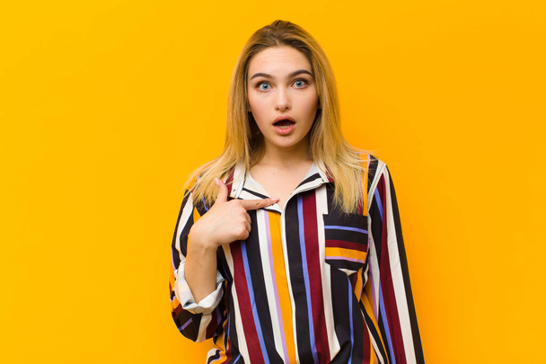 young blonde pretty woman looking shocked and surprised with mouth wide open, pointing to self against orange wall - Foto, afbeelding