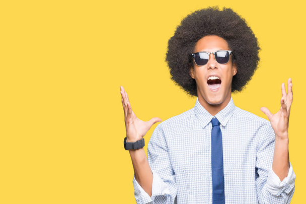 Młody African American Business Man z Afro włosy ubrany okulary przeciwsłoneczne szalony i szalony krzycząc i krzyczeć z agresywnym ekspresji i ramiona podniesione. Koncepcja frustracji. - Zdjęcie, obraz