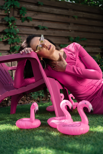 attractive woman in dress and sunglasses posing and sitting on grass outside  - 写真・画像