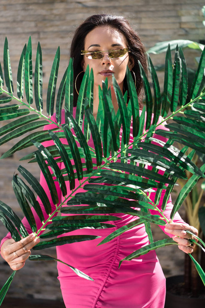 atraktivní žena v šatech a slunečních brýlích pózuje s listy venku  - Fotografie, Obrázek