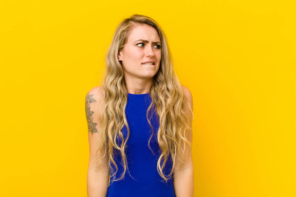 young blonde woman looking worried, stressed, anxious and scared, panicking and clenching teeth against yellow wall - Foto, afbeelding