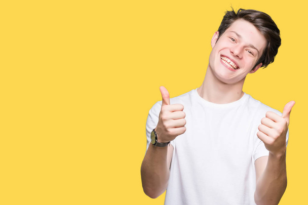 Jonge knappe man dragen casual wit t-shirt over geïsoleerde achtergrond succes teken doen positief gebaar met hand, thumbs up glimlachen en gelukkig. Kijken naar de camera met vrolijke expressie, winnaar gebaar. - Foto, afbeelding