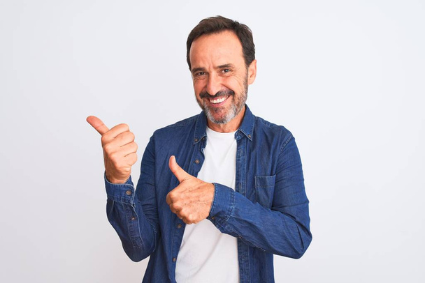 Pohledný muž středního věku v modré džínové košili stojící nad izolovaným bílým pozadím ukazující na zadní stranu s rukou a palci nahoru, s úsměvem sebevědomý - Fotografie, Obrázek