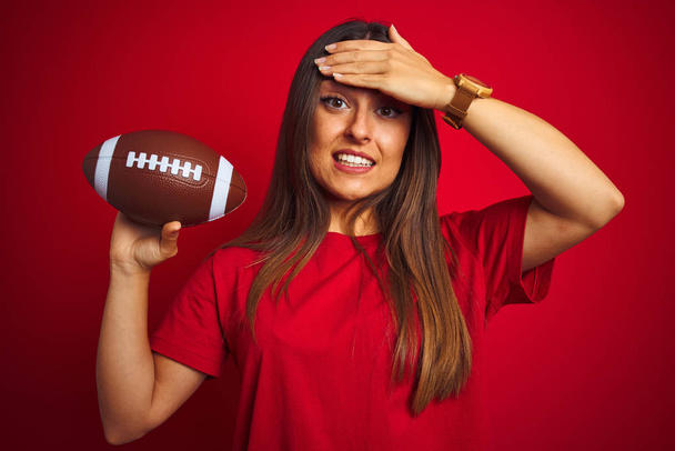 Genç ve güzel sporcu kadın futbol topunu izole edilmiş kırmızı arka planda tutuyor. Elleri başının üstünde, utanç ve şaşkınlık dolu, sinirli ve hayal kırıklığına uğramış bir halde. Hata için korku ve üzüntü. - Fotoğraf, Görsel