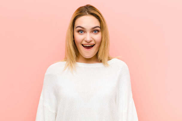 young pretty blonde woman looking happy and pleasantly surprised, excited with a fascinated and shocked expression against pink flat wall - Fotoğraf, Görsel