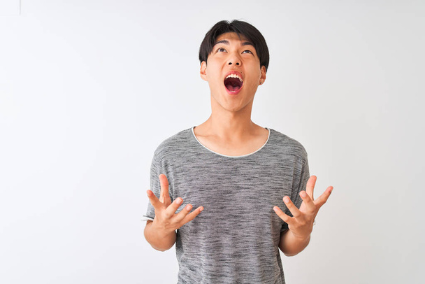 Jonge Chinese man draagt casual t-shirt staan over geïsoleerde witte achtergrond gek en gek schreeuwen en schreeuwen met agressieve uitdrukking en armen omhoog. Frustratieconcept. - Foto, afbeelding