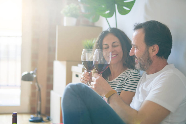 Orta yaş Senior romantik çift aşk yeni bir eve taşımak için mutlu gülümseyen bir bardak şarap içme kutluyor, etrafında kutuları ile daire katında oturan - Fotoğraf, Görsel