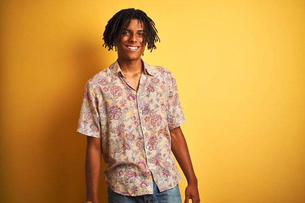 Afro homem com dreadlocks em férias vestindo camisa floral sobre fundo amarelo isolado olhando para o lado com sorriso no rosto, expressão natural. Rindo confiante
. - Foto, Imagem