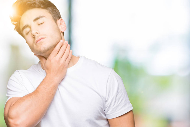 Jonge knappe man dragen witte t-shirt over achtergrond geïsoleerd raken van pijnlijke nek, zere keel voor griep, clod en infectie - Foto, afbeelding