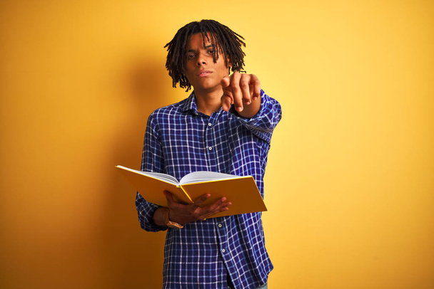 Afro amerykański student człowiek z dredami czytania książki na odizolowanym żółtym tle wskazując palcem na aparat i do Ciebie, znak ręczny, pozytywny i pewny gest z przodu - Zdjęcie, obraz