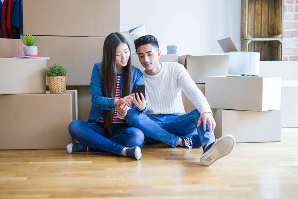 Joven pareja asiática sentada en el suelo de un nuevo apartamento arround cajas de cartón, usando smartphone y sonriendo en un nuevo hogar
 - Foto, imagen