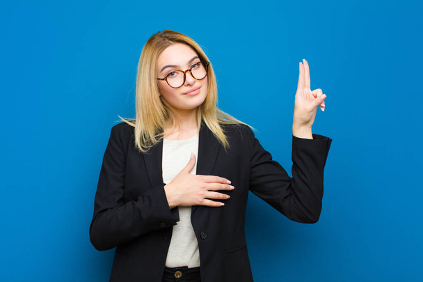 young pretty blonde woman looking happy, confident and trustworthy, smiling and showing victory sign, with a positive attitude against flat wall - 写真・画像