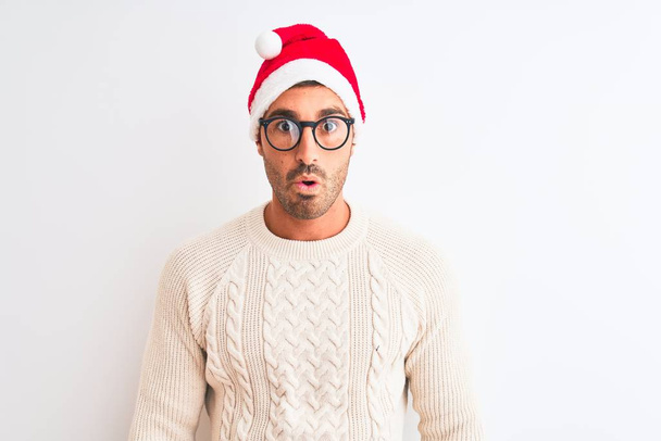 Jonge knappe man draagt kerst hoed en bril over geïsoleerde achtergrond bang en geschokt met verrassende uitdrukking, angst en opgewonden gezicht. - Foto, afbeelding