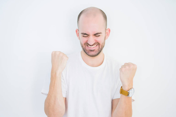 Young bald man over white isolated background very happy and excited doing winner gesture with arms raised, smiling and screaming for success. Celebration concept. - Foto, Imagen
