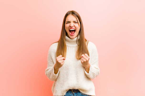 young pretty woman shouting aggressively with annoyed, frustrated, angry look and tight fists, feeling furious against pink background - Fotoğraf, Görsel