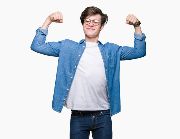 Jeune bel homme portant des lunettes sur fond isolé montrant les muscles des bras souriant fier. Concept de fitness
. - Photo, image
