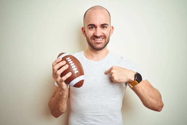 Молодой человек держит регби американский футбольный мяч на изолированном фоне с неожиданным лицом, указывающим пальцем на себя
 - Фото, изображение