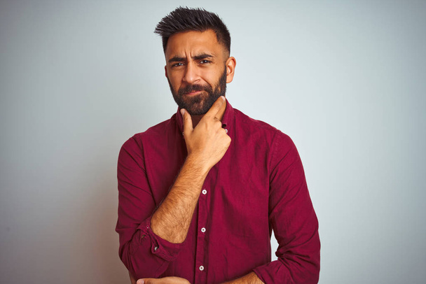 Young indian man wearing red elegant shirt standing over isolated grey background looking confident at the camera smiling with crossed arms and hand raised on chin. Thinking positive. - Zdjęcie, obraz