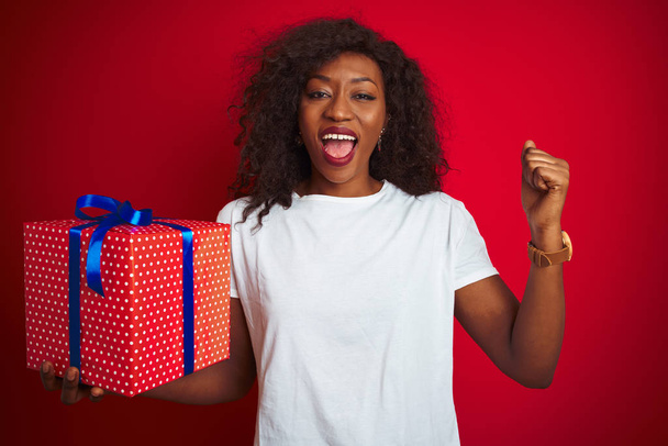 Genç Afrikalı Amerikalı kadın elinde doğum günü hediyesi ile kırmızı arka planda durmuş gururla bağırıyor ve zaferi ve başarıyı kutluyor. - Fotoğraf, Görsel