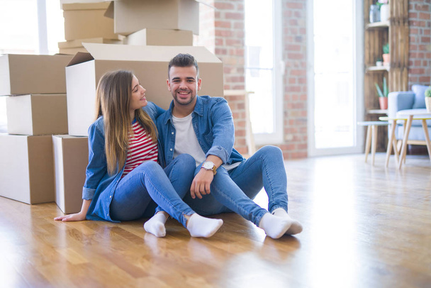 Young beautiful couple in love moving to new home, sitting on the floor very happy and cheerful for new apartment around cardboard boxes - Photo, Image