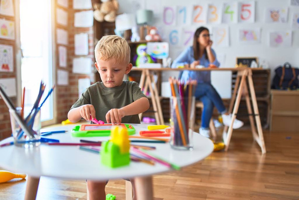 Nuori valkoihoinen lapsi leikkii leikkikoulussa opettajan kanssa. Äiti ja poika leikkihuoneessa piirustus piirtää väri kyniä, nuori nainen taustalla istuu pöydällä
. - Valokuva, kuva