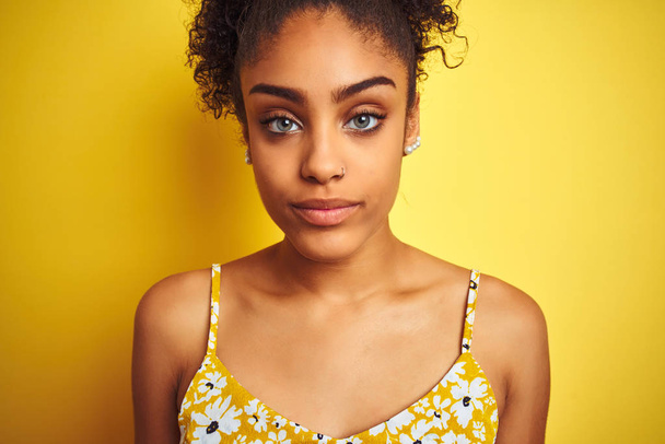 Jovem afro-americana vestindo vestido floral verão sobre fundo amarelo isolado com uma expressão confiante no rosto inteligente pensando sério
 - Foto, Imagem