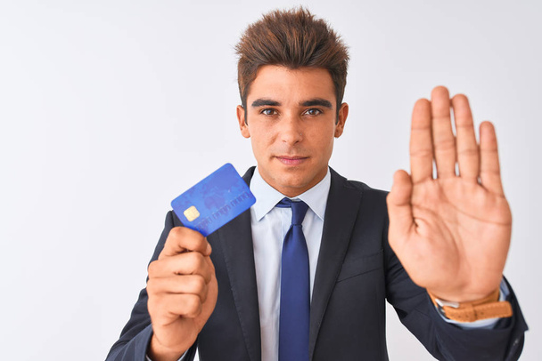 Fiatal jóképű üzletember visel öltöny tartja hitelkártya felett elszigetelt fehér háttér nyitott kézzel csinál stop jel komoly és magabiztos kifejezés, védelmi gesztus - Fotó, kép
