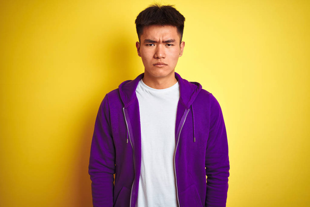 若いアジア系中国人の男性が紫色のスウェットシャツを着て孤立した黄色の背景懐疑的で緊張の上に立って、問題のために動揺を眉をひそめている。負の人. - 写真・画像