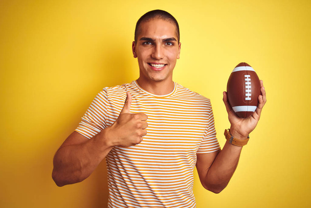 junger Rugbyspieler Mann hält einen Fußball über gelbem isolierten Hintergrund glücklich mit breitem Lächeln tut ok Zeichen, Daumen hoch mit den Fingern, ausgezeichnetes Zeichen - Foto, Bild