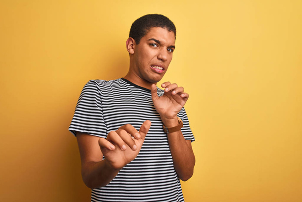 Young handsome arab man wearing navy striped t-shirt over isolated yellow background disgusted expression, displeased and fearful doing disgust face because aversion reaction. With hands raised. Annoying concept. - Photo, Image