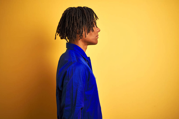 Afro worker man with dreadlocks wearing mechanic uniform over isolated yellow background looking to side, relax profile pose with natural face with confident smile. - Фото, зображення