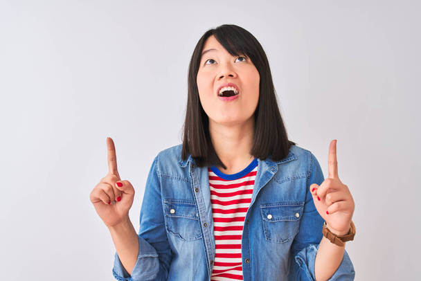 Mujer china con camisa de mezclilla y camiseta de rayas rojas sobre fondo blanco aislado asombrada y sorprendida mirando hacia arriba y señalando con los dedos y los brazos levantados
. - Foto, imagen