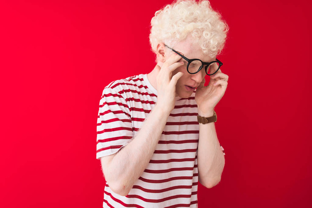 Mladý albín blond muž nosí pruhované tričko a brýle přes izolované červené pozadí s rukou na hlavě pro bolest hlavy, protože stres. Trpící migréna. - Fotografie, Obrázek