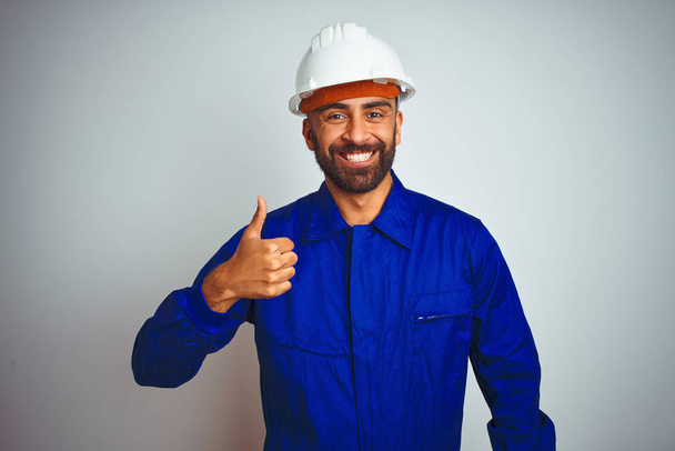 Beau travailleur indien portant uniforme et casque sur fond blanc isolé faisant des pouces heureux geste avec la main. Approuver l'expression en regardant la caméra montrant le succès
. - Photo, image