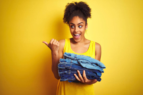jonge afrikaanse amerikaanse vrouw houden stapel jeans over geïsoleerde gele achtergrond wijzen en tonen met duim naar de zijkant met gelukkig gezicht glimlachen - Foto, afbeelding