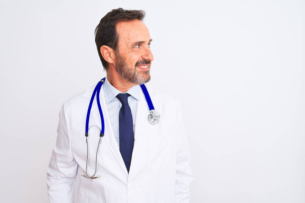 Μεσήλικας γιατρός άνδρας φορώντας παλτό και στηθοσκόπιο στέκεται πάνω από απομονωμένο λευκό φόντο κοιτάζοντας μακριά στην πλευρά με χαμόγελο στο πρόσωπο, φυσική έκφραση. Γελάς σίγουρος. - Φωτογραφία, εικόνα