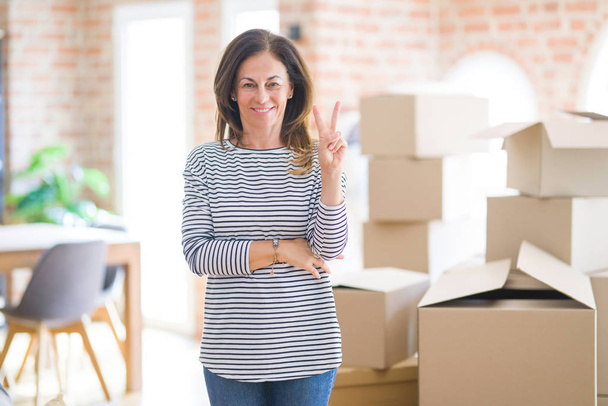 Middle age woman moving to a new house arround cardboard boxes smiling with happy face winking at the camera doing victory sign. Number two. - Photo, Image