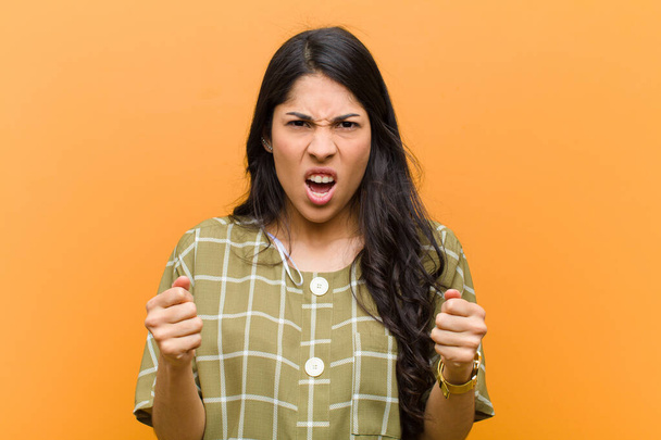 young pretty hispanic woman shouting aggressively with annoyed, frustrated, angry look and tight fists, feeling furious against brown wall - Fotoğraf, Görsel