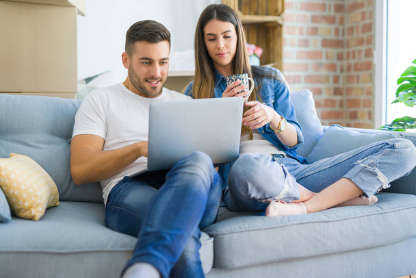 若いカップルは、コンピュータのラップトップを使用してソファに座ってリラックスして新しい家に移動し、新しいアパートに引っ越すために幸せに笑顔 - 写真・画像