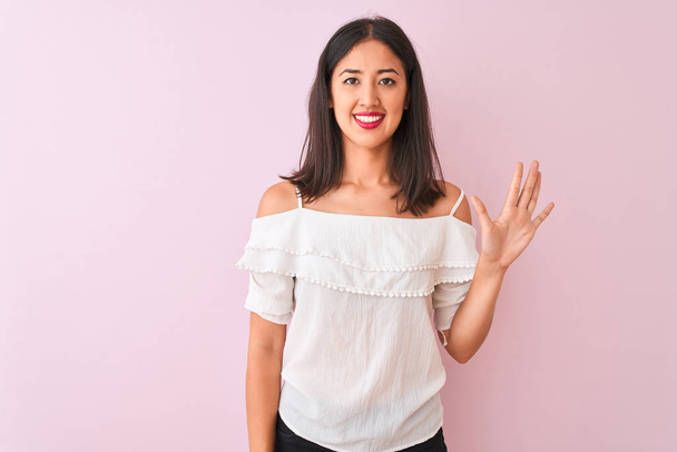 Красивая китаянка в белой футболке стоит на изолированном розовом фоне и показывает пальцами номер пять, улыбаясь уверенно и счастливо
. - Фото, изображение