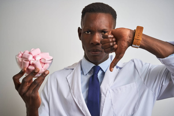 afrikanisch-amerikanischer Arzt hält Schale mit Marshmallow über isoliertem weißem Hintergrund mit wütendem Gesicht, Negativzeichen zeigt Abneigung mit gedrückten Daumen, Ablehnungskonzept - Foto, Bild